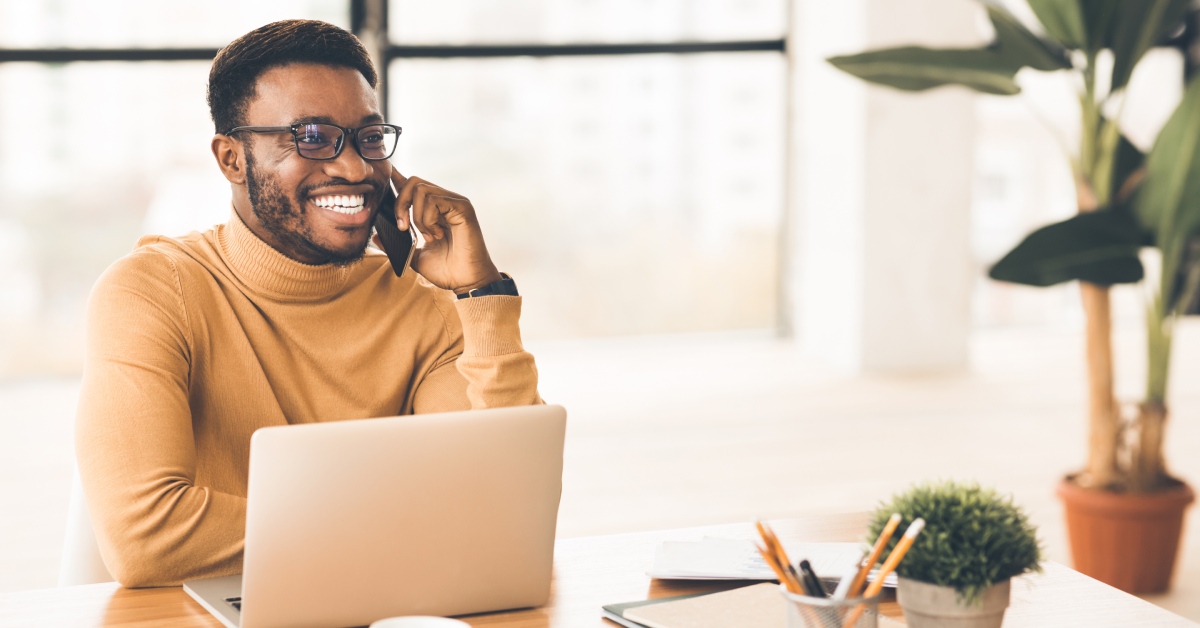 Someone wearing a tan turtle neck is smiling while sitting at their desk and talking on their cell phone.