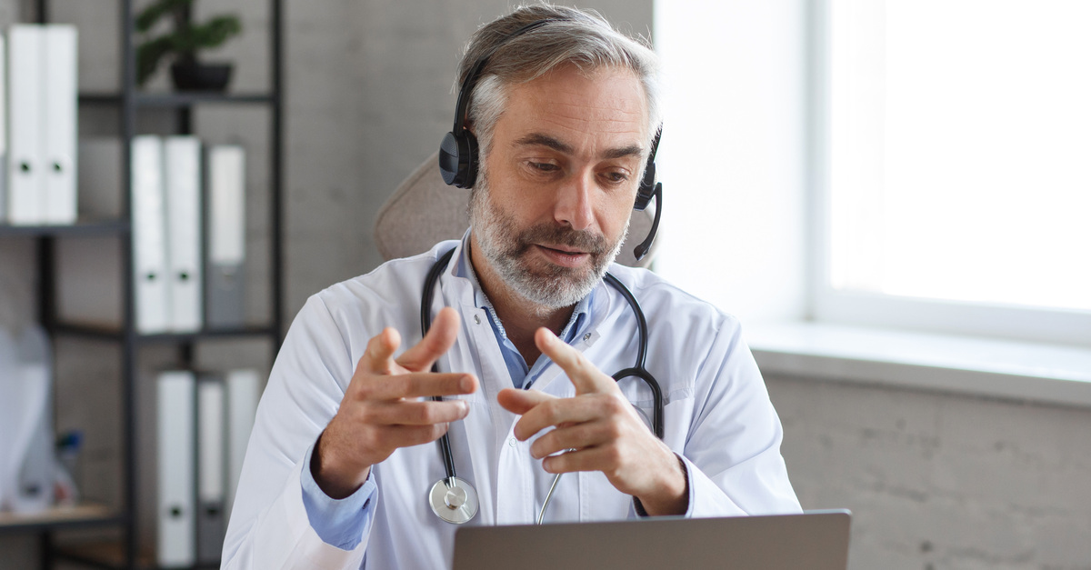 A doctor is wearing a headset and sitting down in front of a laptop. The doctor is on a video call and pointing.