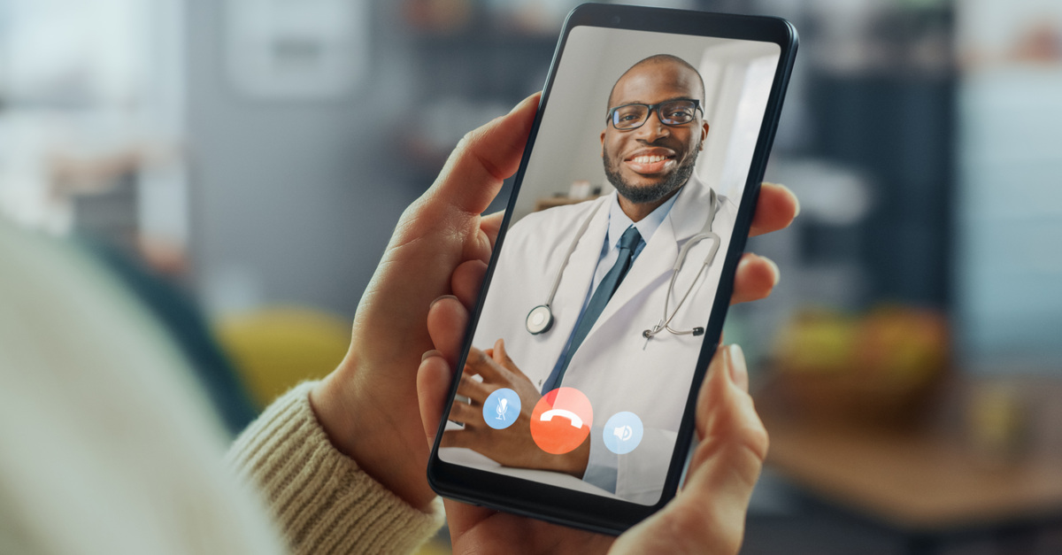 A person is holding a cell phone in their hands while having a video call with a doctor wearing glasses.