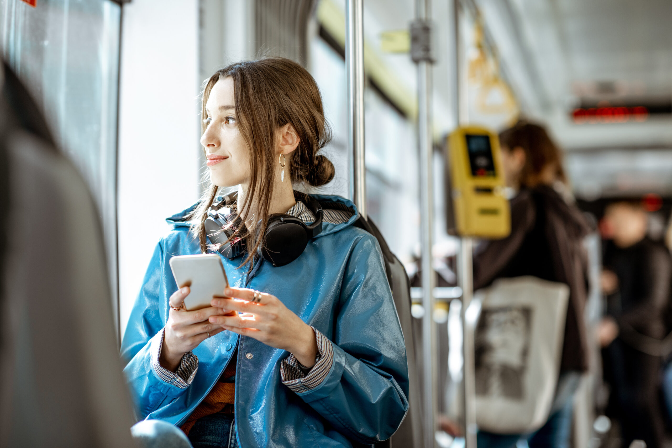 A person is wearing a blue jacket and headphones while holding their cell phone and sitting on a train.