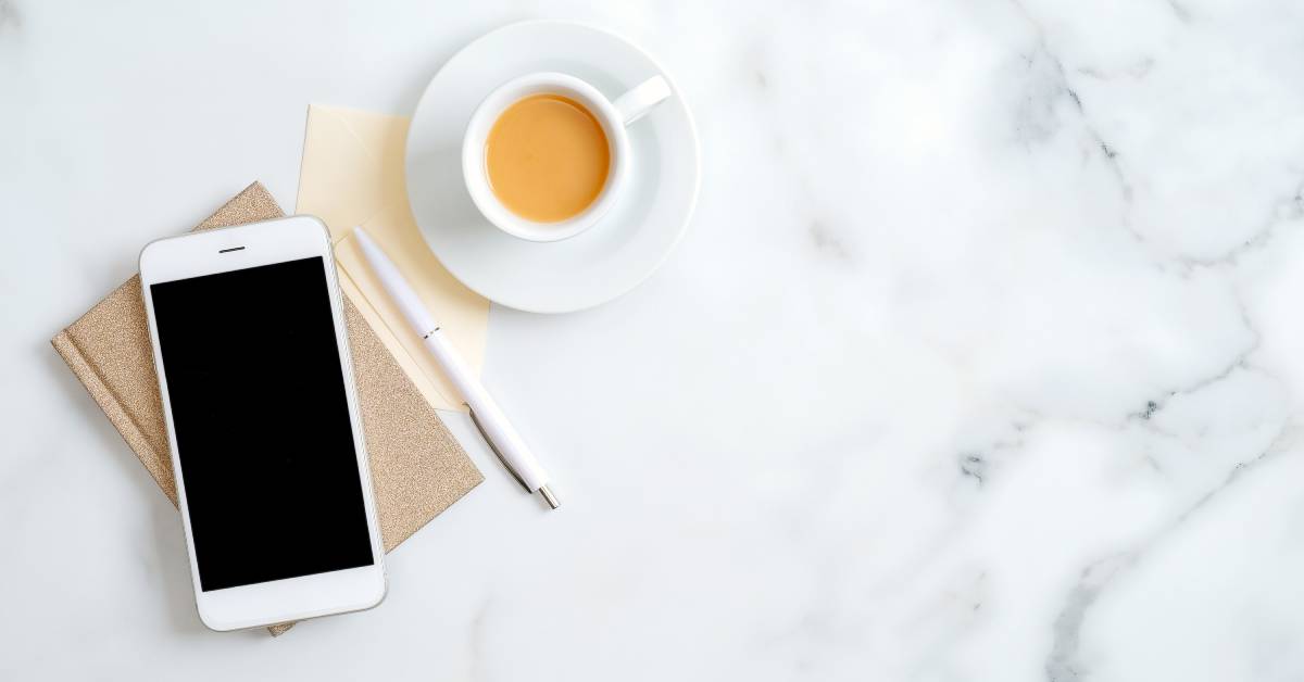 A white cell phone sitting on a brown notebook next to a white pen and a white cup that is full of coffee.