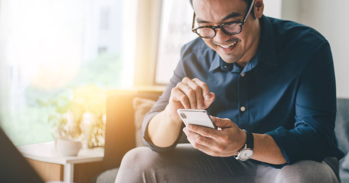 A person wearing a dark blue shirt and brown glasses. They smile and stare at their cell phone screen.