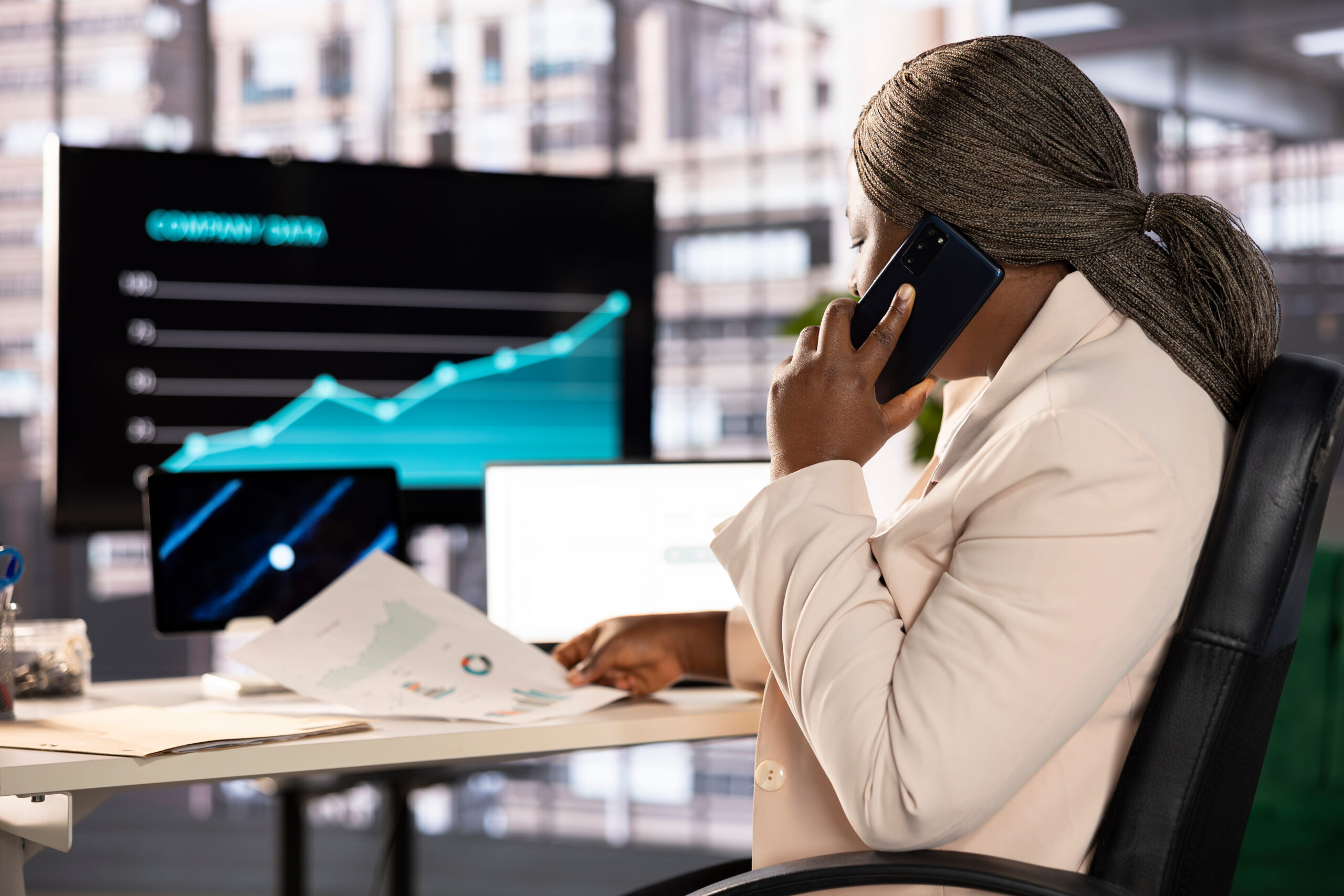 A worker is sitting at their desk while talking on their cell phone. The desk has three different monitors on it.