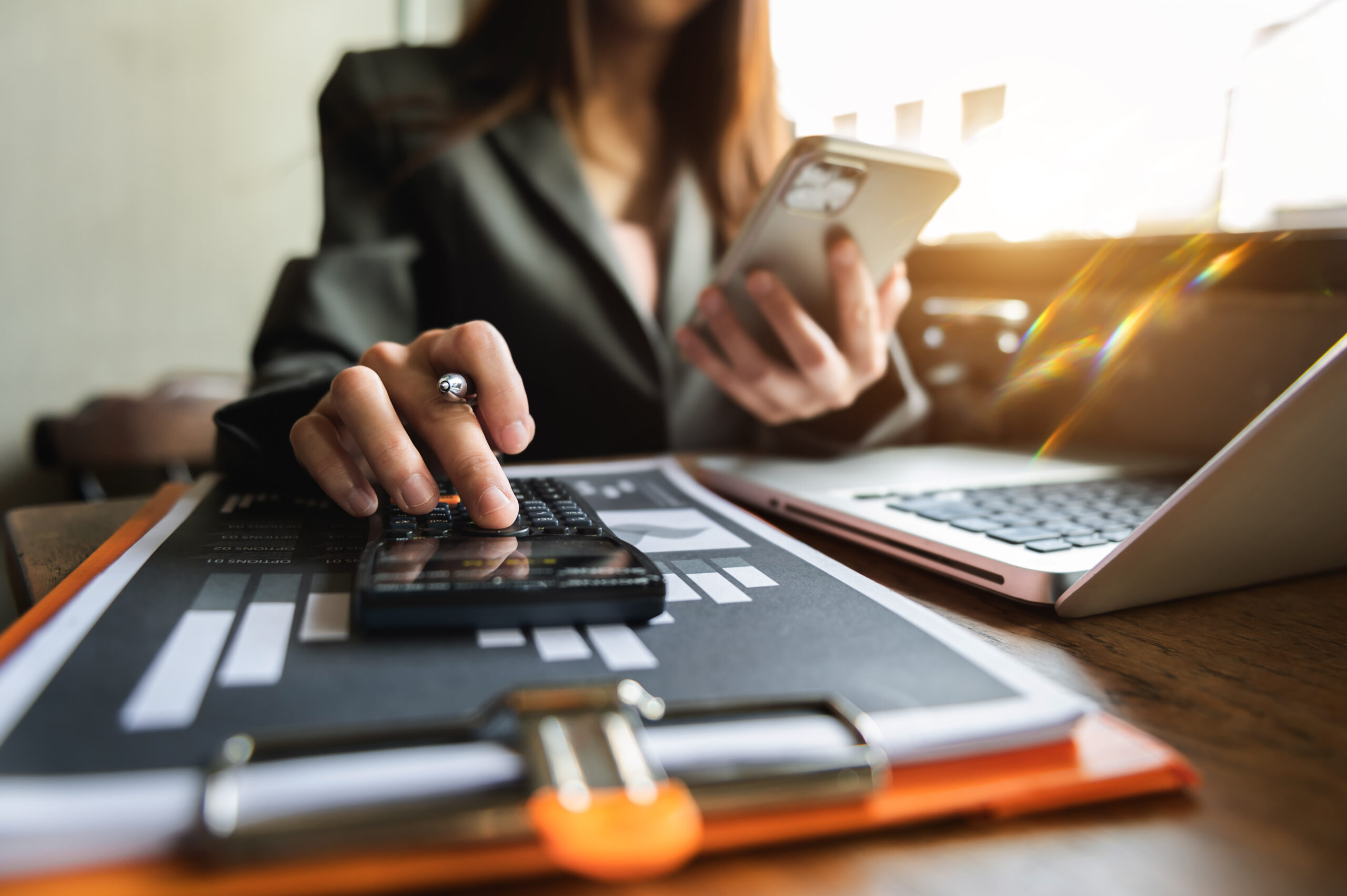 A person wearing a business suit is holding a cell phone while tapping the buttons on a black calculator.