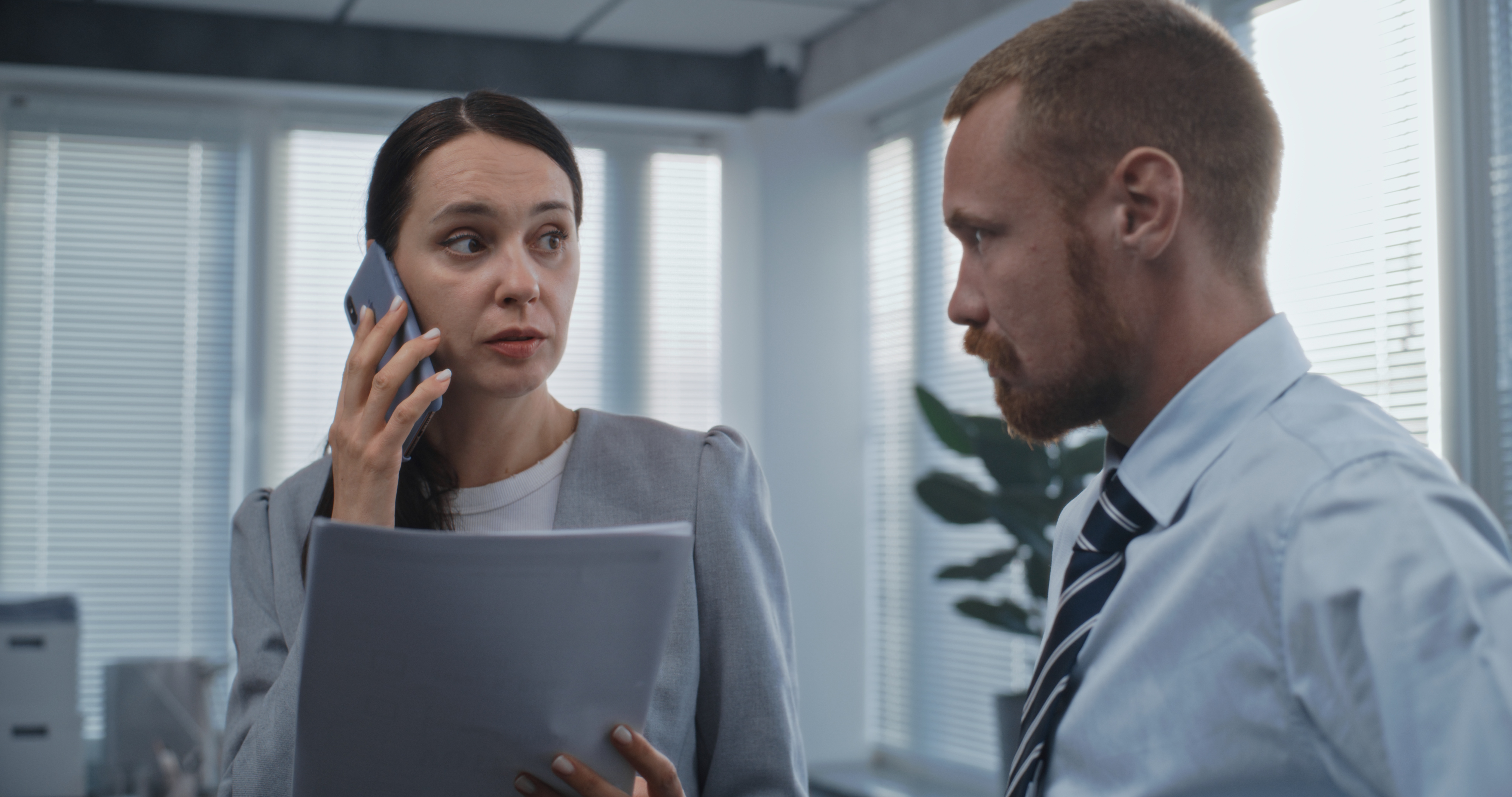 A person holding documents and a cell phone looks at a person to their left. They are both wearing business suits.