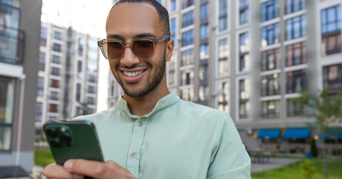 Someone wearing a green shirt is holding a green cellphone. They are standing outside surrounded by tall buildings.