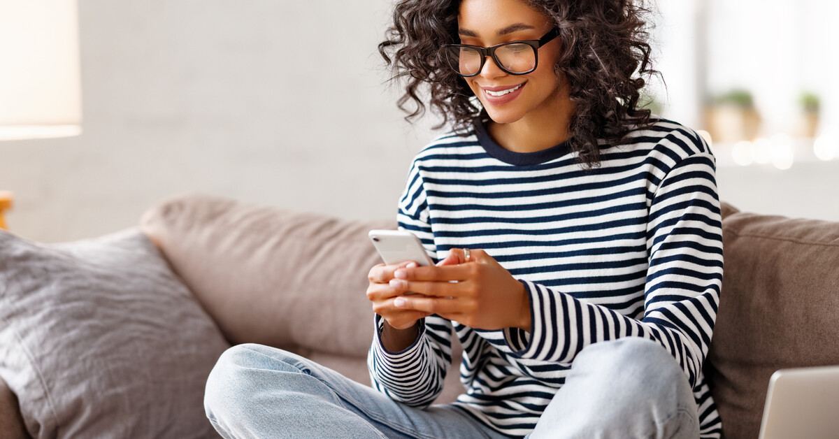 A person wearing glasses and a striped long-sleeve shirt smiles while sitting on a couch and texting on their cellphone.