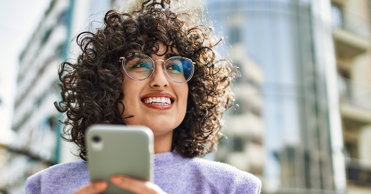 A person wearing glasses and a purple long-sleeve shirt is smiling and texting outside in front of a tall building.