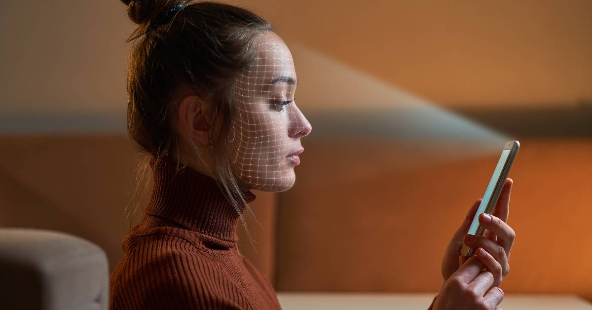 A person wearing a turtleneck is sitting by an orange wall and using facial scanning technology on their cell phone.