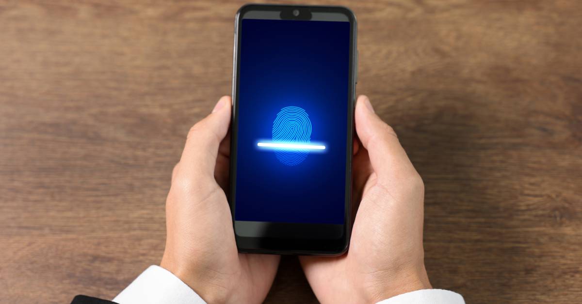 A person is holding a cell phone in their hands over a wooden table. The phone screen has a fingerprint scanner on it.