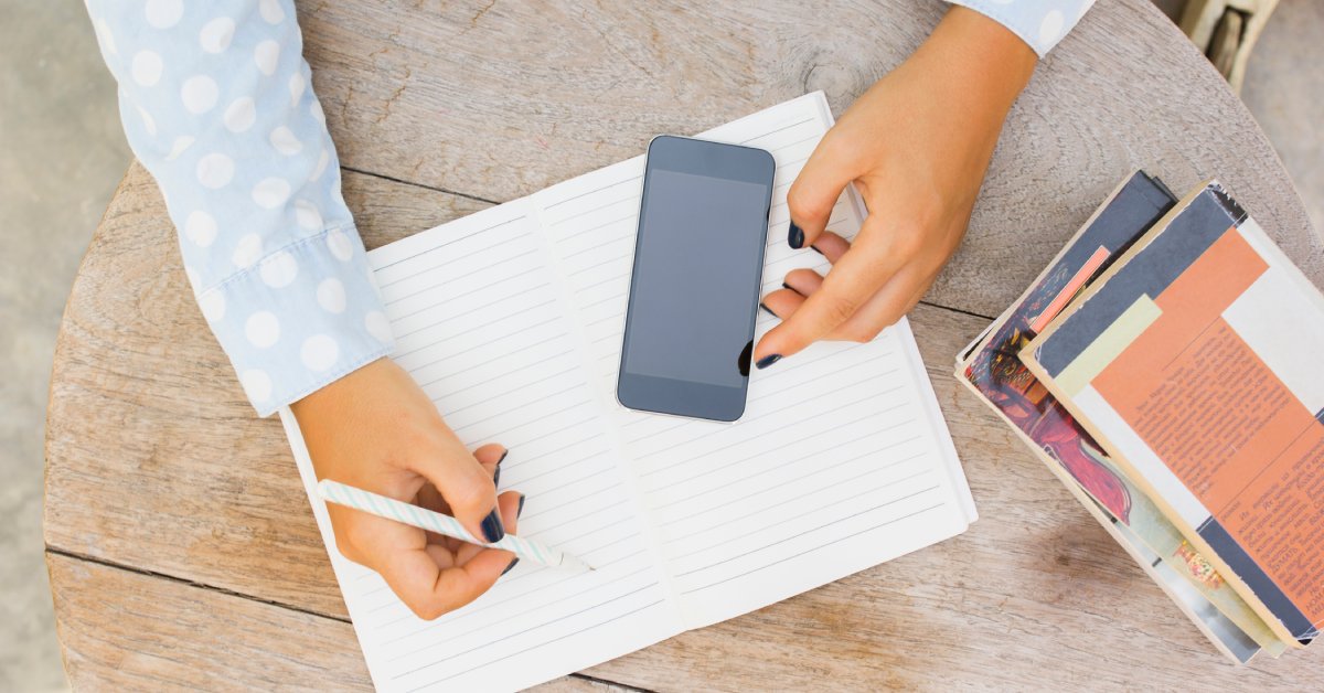 A person is holding a striped pen over an open notebook. A cell phone is lying on top of the notebook.