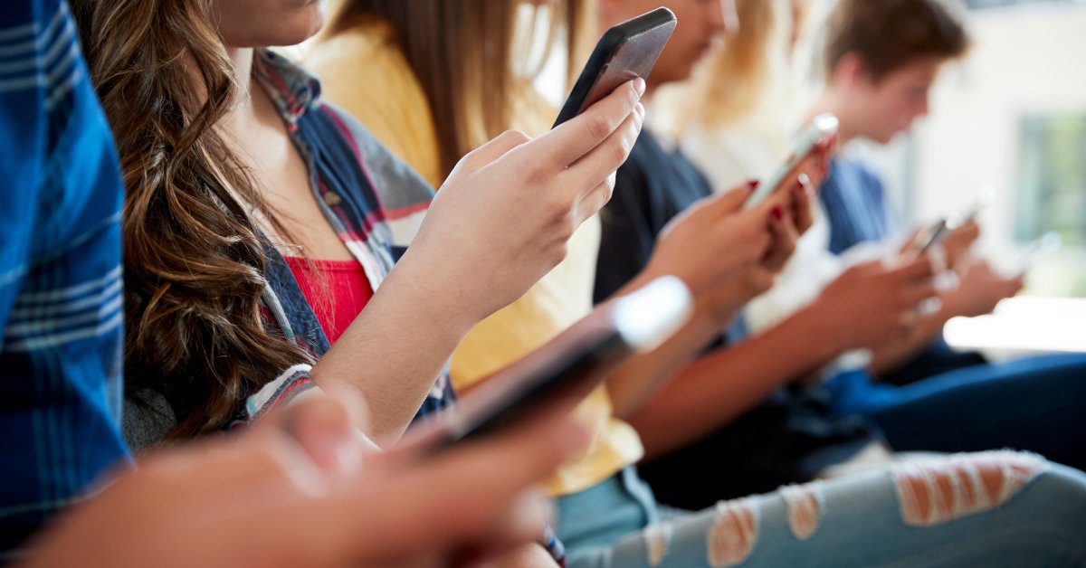 A group of six kids are sitting down during the day and looking down at the cell phones in their hands.