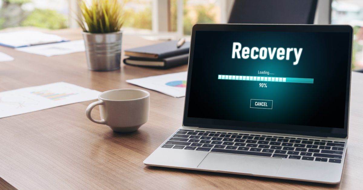 A laptop with “Recovery” labeled on the screen above a loading bar that is at 90%. The laptop is on a wooden desk.