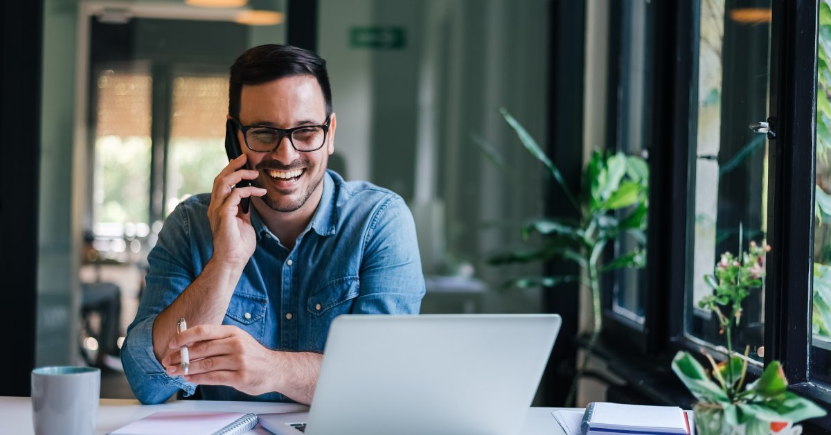 A person talks on a cell phone and sits at a desk. They have a phone in their right hand and a pen in their left hand.