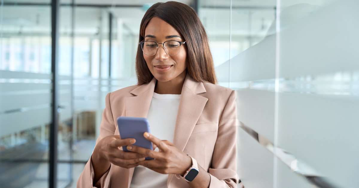 A business professional holds a purple cell phone. They are looking down at the phone while leaning on a window.