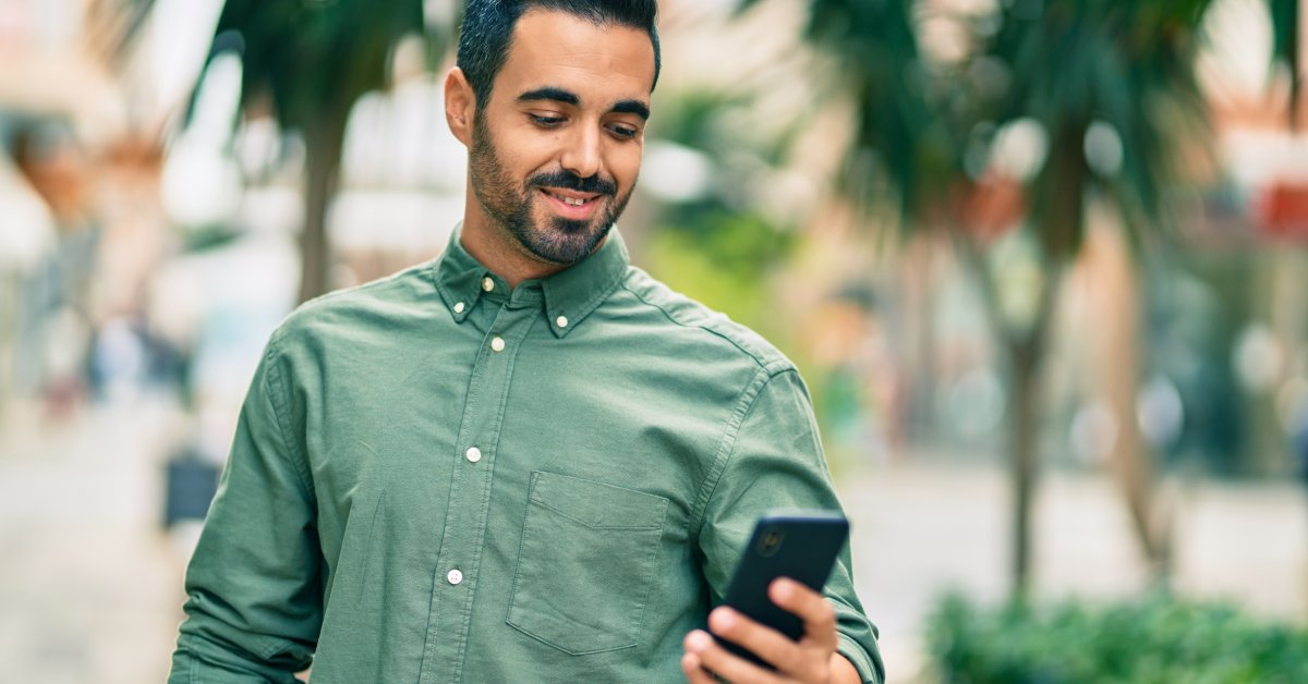 A person wears a green shirt while standing next to trees outside. They are smiling and looking at their cell phone.