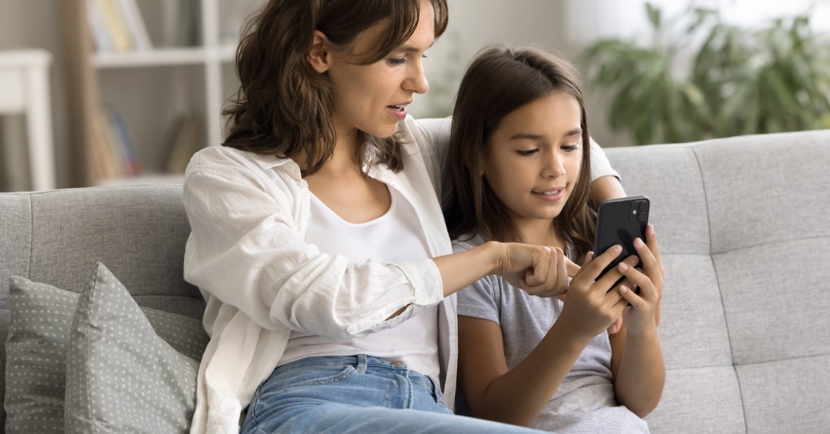 A parent sitting with their child on a gray couch. The parent is pointing at a phone the child is holding.