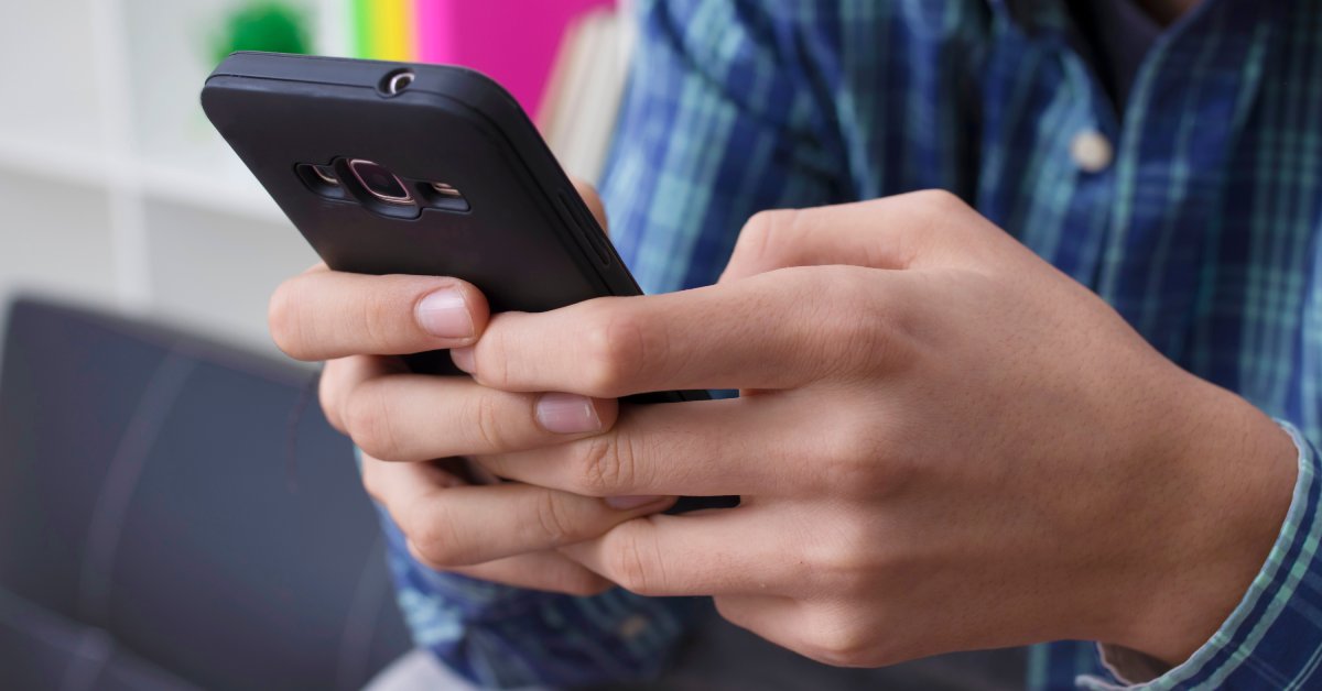 A child is wearing a blue shirt and holding a cell phone with a black protective cover in their hands.