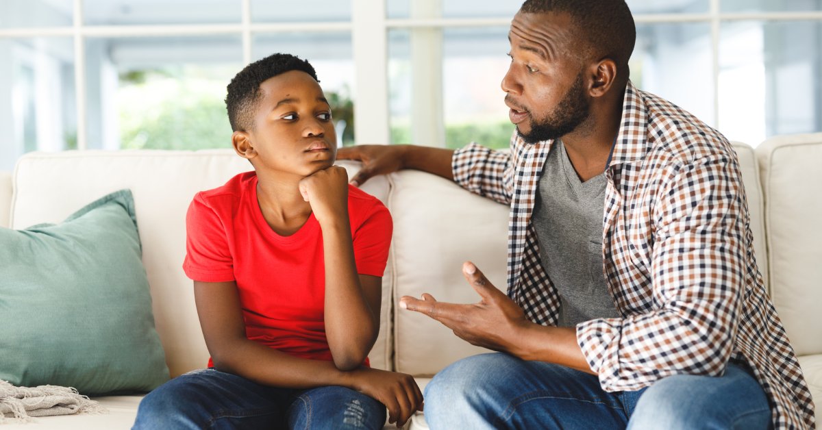 A parent talks to their child while sitting on a white couch with a green pillow. Large windows are in the background.