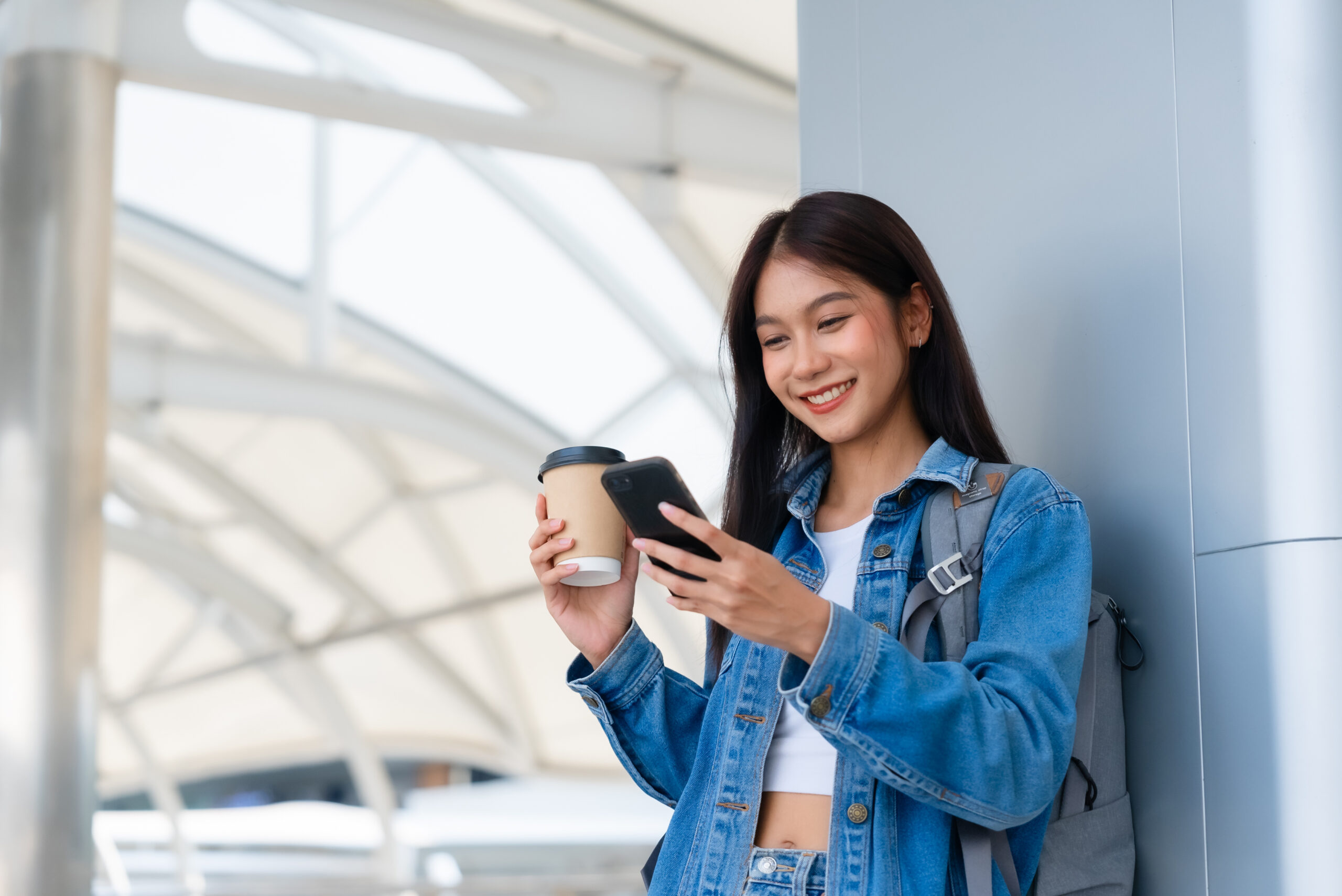 A person wearing a backpack is standing in a large public space while holding a coffee cup and a phone.