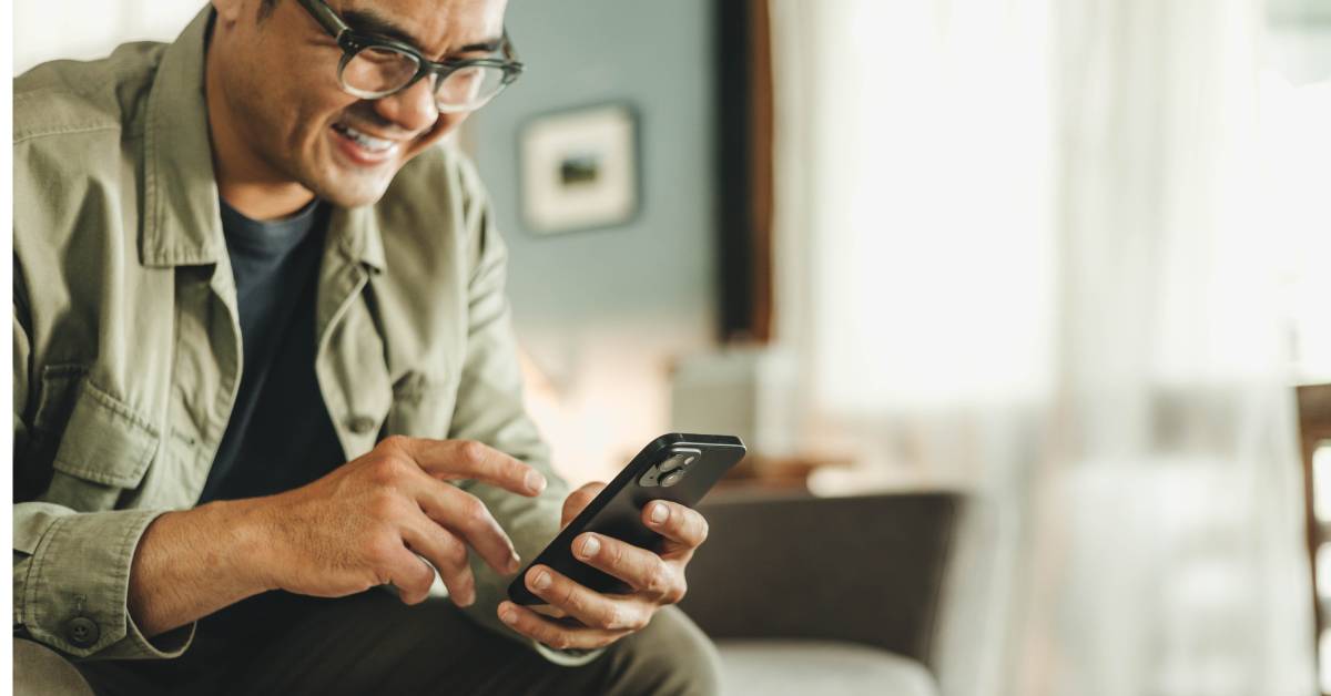 A person wearing glasses and a jacket is chuckling as they move their right hand toward their cell phone.