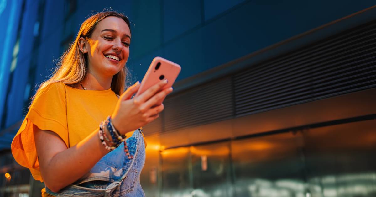 A person wearing blue overalls and a yellow shirt smiles while walking down the street and looking at their phone.