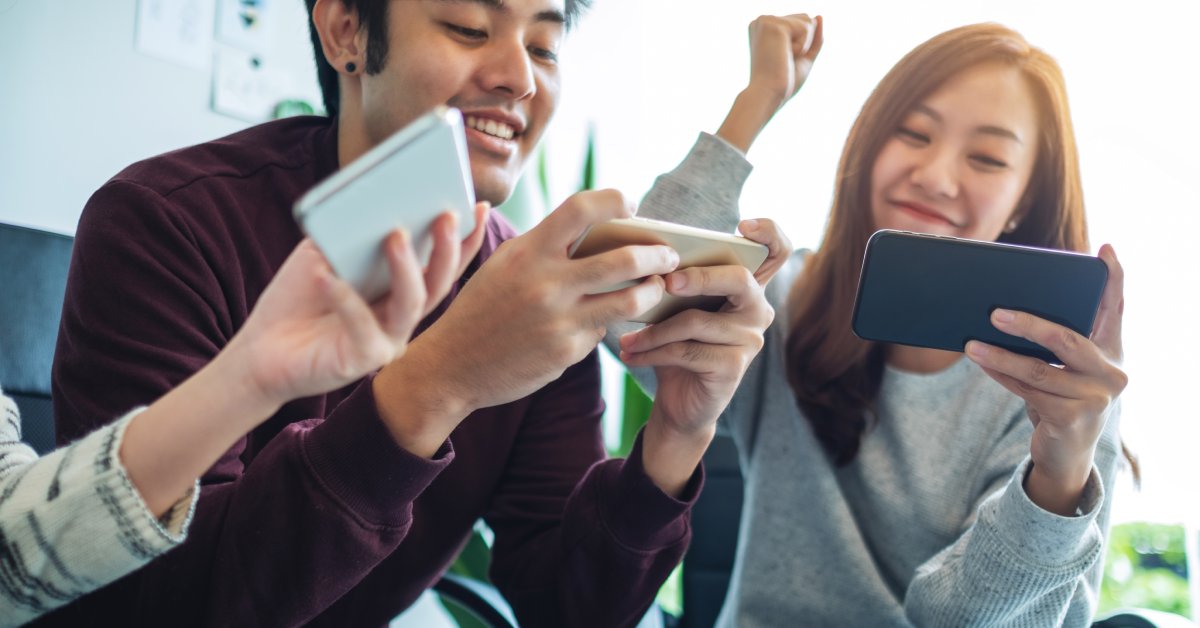 Three people are sitting outside and playing video games on their cell phones. One person pumps their fist into the air.