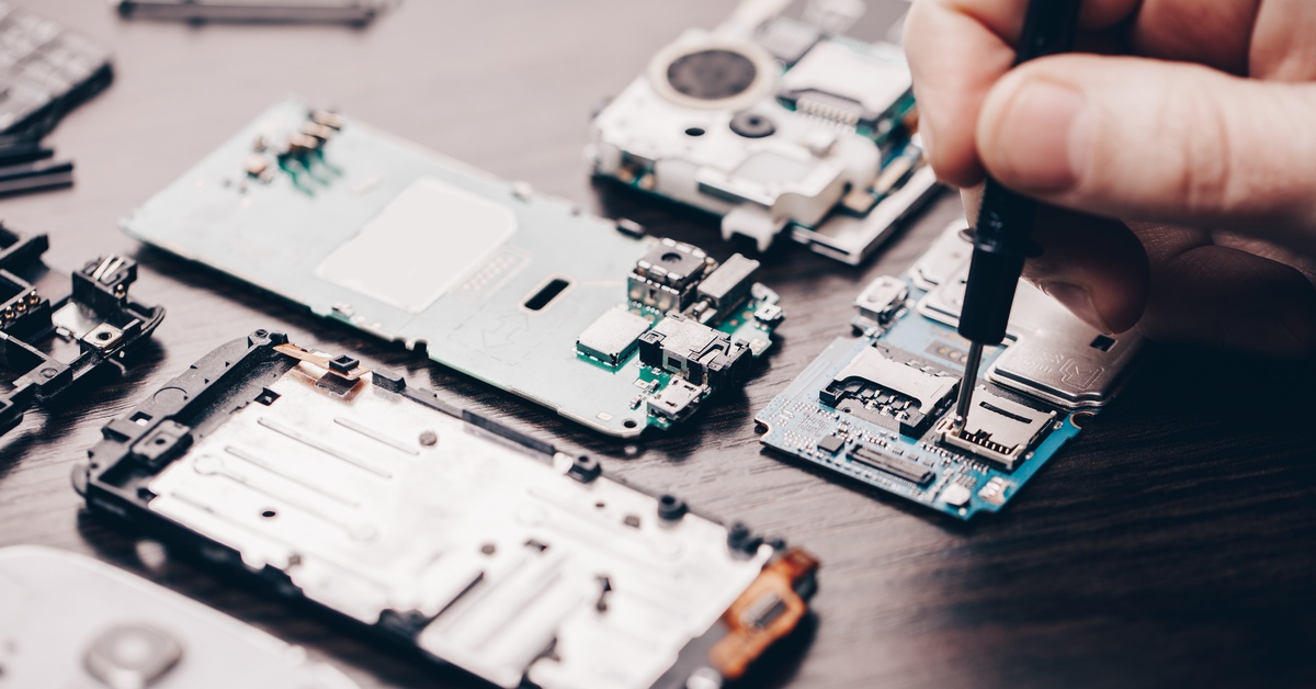 A cell phone is dismantled on a brown table. Someone is repairing the phone with a small black tool.