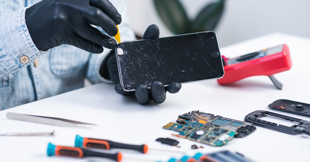 A repair specialist wearing work goggles is holding a dismantled cell phone in front of a full workbench.