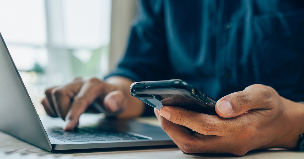 A person is sitting down at a desk indoors. They are holding a cell phone and tapping the keyboard on their laptop.