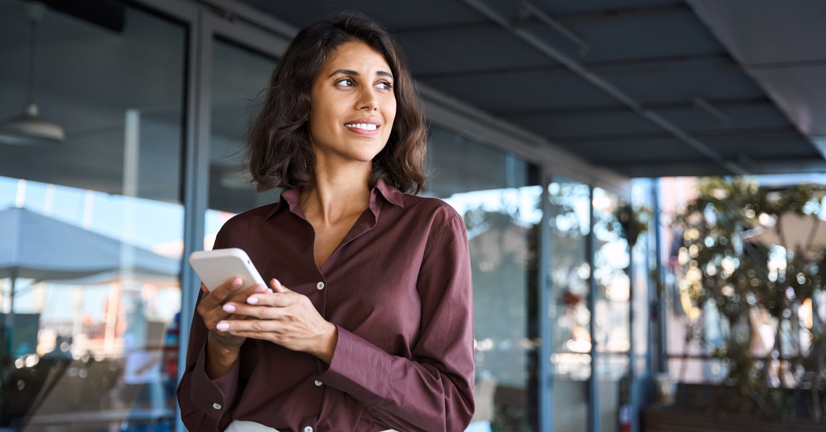 A professionally dressed person is holding their white cell phone and standing outside during the day.