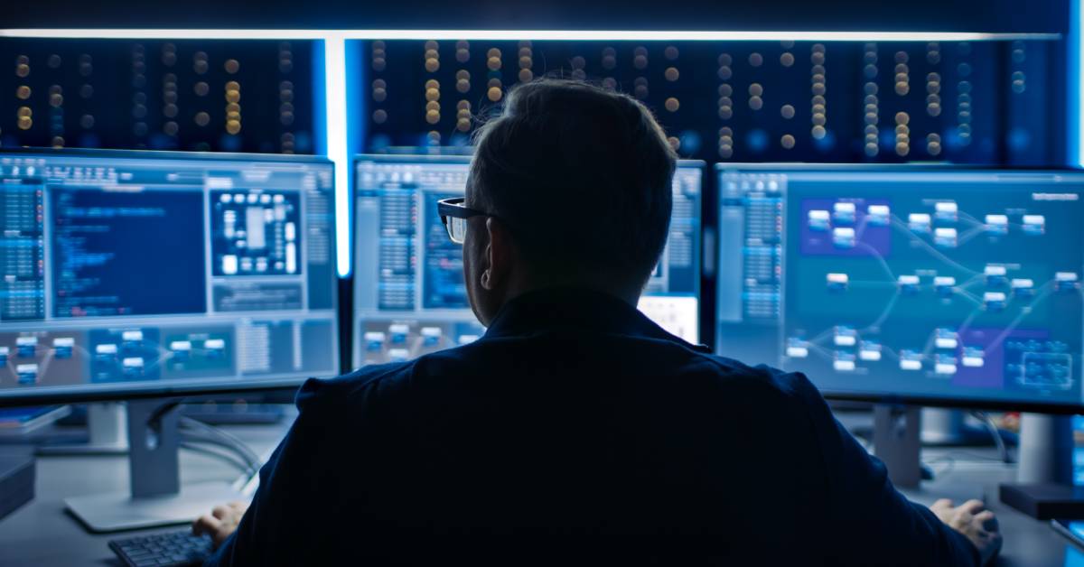 A person wearing glasses sits down at a desk. They are sitting in front of three computer monitors.