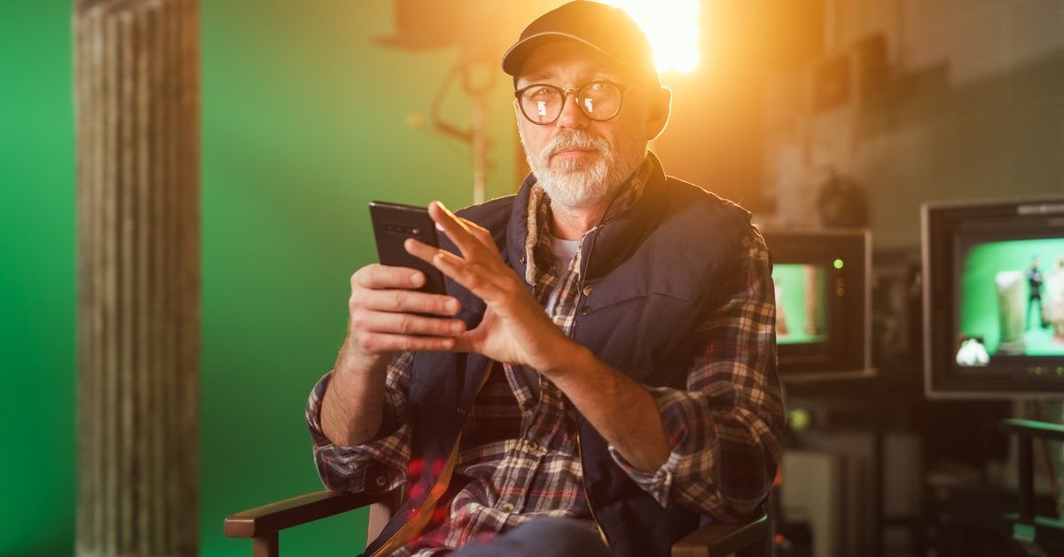 A person wearing a hat and glasses sits in a director’s chair on a film set. They are holding a phone in their hands.